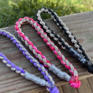 Op de afbeelding: Drie gevlochten koorden met een herhalend patroon van gekleurd touw en zilverkleurige strass-steentjes. De koorden zijn paars, roze en zwart, elk met een grijs rubberen label met de tekst Maddie Jade en een klein logo-icoontje.