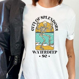 May include: White t-shirt with a graphic of a skeleton holding two beers and standing on a beach with the text "City of Splendors Waterdeep .sc."