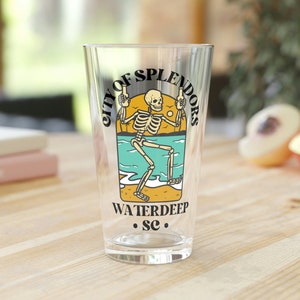 May include: A clear glass pint glass with a graphic of a skeleton holding two drinks on a beach with the text "City of Splendors Waterdeep SC".