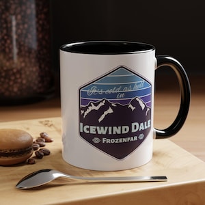 May include: White ceramic mug with a black rim and handle. The mug features a blue and purple mountain range graphic with the text "It's cold as hell in Icewind Dale FROZENFAR".