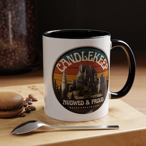 May include: White ceramic mug with a black interior and handle. The mug features a circular design with the text "CANDLEKEEP" and "AVOWED & PROUD" over a castle illustration with a sunset background. A spoon and a macaron are on the wooden surface.