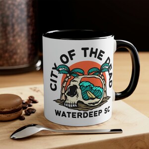 Op de afbeelding: Witte keramische mok met een zwarte binnenkant en handvat. De mok heeft een afbeelding van een schedel, palmbomen en oceaangolven met de tekst "CITY OF THE DEAD WATERDEEP SC". De mok staat op een houten oppervlak.