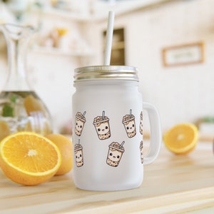 Op de afbeelding: Een witte mason jar met een zilveren deksel en handvat, met een herhalend patroon van schattige cartoon boba thee bekers. De pot heeft een wit rietje en staat op een houten oppervlak met sinaasappelschijfjes.