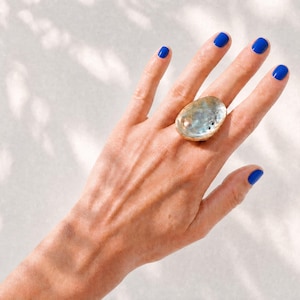 Könnte beinhalten: Ein Ring mit einem großen, ovalen Cabochon aus irisierendem Abalone-Muschelgehäuse, gefasst in ein goldfarbenes Metallband. Die Muschel zeigt ein natürliches, wirbelndes Muster aus Blau-, Grün- und Silbertönen mit einer polierten Oberfläche.
