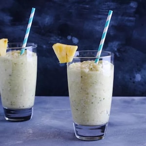 Puede incluir: Dos vasos altos llenos de una bebida verde claro, adornados con trozos de piña y pajitas a rayas. Las bebidas están sobre una superficie azul contra un fondo azul oscuro, lo que sugiere una bebida refrescante.