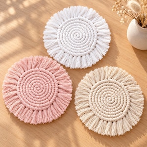 Könnte beinhalten: Drei kreisförmige Makramee-Untersetzer in Weiß, zartem Rosa und Beige. Jeder Untersetzer weist ein zentrales Spiralmuster aus geknoteter Kordel auf, das von einem Fransenrand aus losen, geraden Strängen umgeben ist.