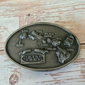 May include: Oval-shaped silver-toned belt buckle featuring a detailed relief map of the Hawaiian Islands. The buckle includes the names of the islands and the word "HAWAII" in a banner.