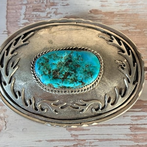 May include: An oval-shaped metal belt buckle featuring a central, polished turquoise stone with dark matrix inclusions. The stone is framed by a twisted rope border, and the surrounding metal surface is decorated with cut-out branch-like patterns.