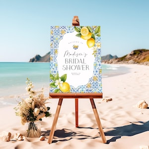 May include: A wooden easel displays a bridal shower sign on a sandy beach. The sign features a blue and white tile pattern with lemons and the text "Madison's BRIDAL SHOWER". A bouquet of flowers sits in a glass vase.