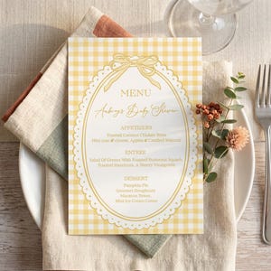 May include: A baby shower menu card with a yellow gingham pattern and an oval frame. The menu includes appetizers, entrees, and desserts. A fork, knife, and plate are set on a linen napkin.