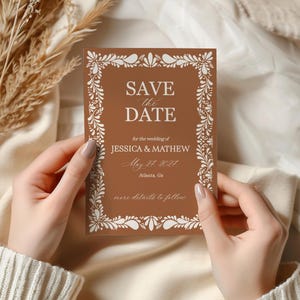 May include: A brown Save the Date card with white floral border and text. The card reads "SAVE the DATE" and includes wedding details for Jessica & Mathew, May 24, 2024, Atlanta, Ga. The card is held by hands.