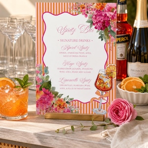 May include: A rectangular Spritz Bar menu sign featuring orange and white vertical stripes, a scalloped white center, and floral illustrations. The text lists signature drinks including Aperol Spritz, Hugo Spritz, and Limoncello Spritz with ingredients.