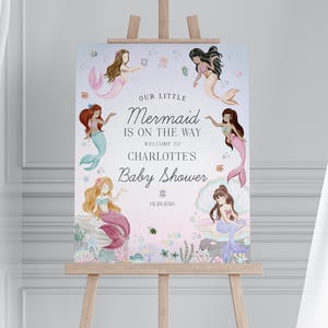May include: A baby shower sign featuring watercolor illustrations of mermaids with flowing hair and colorful tails. The sign reads "Our Little Mermaid is on the Way" and welcomes guests to Charlotte's baby shower, with the date 12.21.2026.