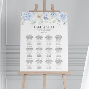 May include: A white wedding seating chart on a wooden easel, featuring floral accents in blue and cream. The text reads "TAKE A SEAT" above the names "RYAN & SOPHIA" and the date "May 6, 2023". The chart lists guest names under table numbers.