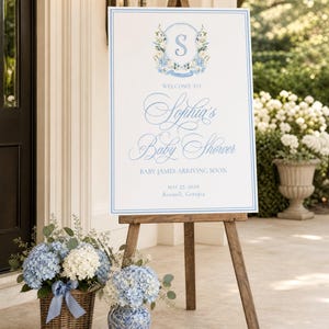 May include: A white rectangular welcome sign on a wooden easel featuring a blue floral crest with the letter S, blue script text reading Welcome to Sophia's Baby Shower, Baby James Arriving Soon, May 27, 2029, and Roswell, Georgia, with blue borders.