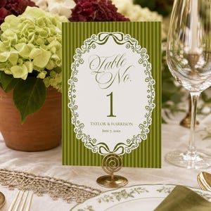 May include: A rectangular table number card featuring a vertical olive green striped background and a white oval frame with floral and ribbon details. The text reads Table No. 1, Taylor & Harrison, and June 7, 20xx, held by a gold spiral stand.