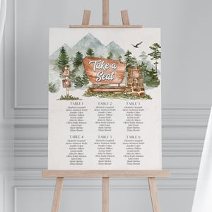 May include: A watercolor-style seating chart displayed on a wooden easel. The chart features a mountain and forest scene with the text "Take a Seat." Six tables are listed with guest names. The overall design has a rustic, outdoorsy theme.