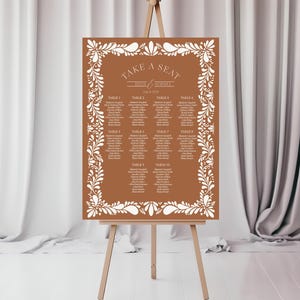 May include: A rectangular wedding seating chart on a wooden easel. The chart has a brown background with white floral border and text. The text reads "Take a Seat" and lists names and table assignments. The easel is made of wood.