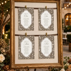 May include: A large, ornate gold-toned frame holds a beige board with two horizontal dark brown ribbons. Four rectangular cards with gray floral patterns and white centers are clipped to the ribbons, displaying table numbers and guest names.