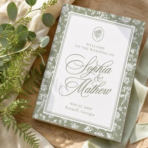 May include: A rectangular wedding invitation card featuring a sage green floral border and a white center panel. The text reads: Welcome to the wedding of Sophia & Mathew, May 27, 2029, Roswell, Georgia. A small floral icon sits above the names.