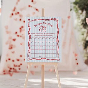 May include: A light blue and white striped sign on a wooden easel. The sign has red text that reads "Guess the Date" and a calendar grid to guess the baby's arrival date. Pink hearts are scattered around the sign.