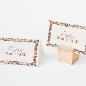 May include: Two custom place cards with brown floral borders and text. One card is propped up by a small wooden block. The cards have the words "Custom PLACE CARD" in a decorative font.