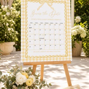 May include: A rectangular sign on a wooden easel featuring a yellow and white gingham border. The text reads Guess the Date, WHEN WILL BABY SOPHIA ARRIVE?, a calendar for September and October, and THE GUEST CLOSEST TO THE ARRIVAL DATE WINS!