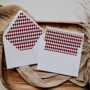 May include: Two white envelopes with a burgundy diamond pattern lining. The envelopes are open, revealing the patterned interior. They rest on a wooden tray with a tan fabric and dried plants.