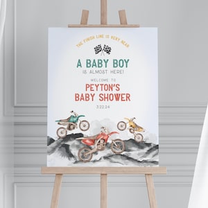 May include: A baby shower sign on an easel. The sign reads "A BABY BOY IS ALMOST HERE!" and welcomes guests to Peyton's baby shower. The sign features watercolor illustrations of three dirt bikes and the date 3.22.24.