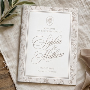 May include: A rectangular wedding program booklet with a beige floral border. The center text reads: Welcome to the wedding of Sophia & Mathew, May 27, 2029, Roswell, Georgia. A small floral icon is centered above the names.