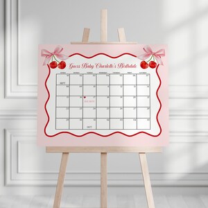 May include: A pink calendar with a red wavy border, displayed on a wooden easel. The calendar features the text "Guess Baby Charlotte's Birthdate" and is decorated with cherry illustrations and pink bows. The calendar grid shows the months of September and October.