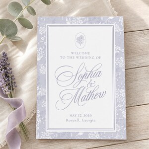 May include: A rectangular wedding invitation card featuring a light purple floral border and a white center panel. The text reads: Welcome to the wedding of Sophia & Mathew, May 27, 2029, Roswell, Georgia. A small floral icon sits above the names.
