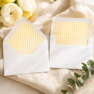 May include: Two white envelopes with yellow gingham patterned interiors are open, displaying the design. The envelopes are on a beige cloth, with yellow flowers and greenery in the background. The envelopes are a stationery item.