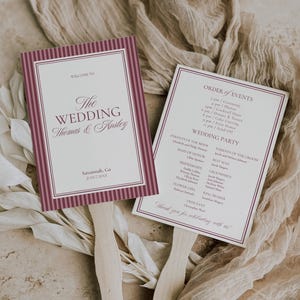 May include: Two wedding fan programs with wooden handles. The fans have a white background with burgundy text and borders. One fan reads "Welcome to The Wedding Thomas & Ansley" and the other lists the order of events and wedding party.