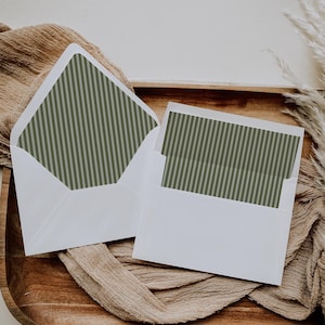 May include: Two white envelopes with green and gray striped interior lining, resting on a wooden tray with a tan cloth. One envelope is open, revealing the striped pattern. The other envelope contains a card with the same striped design.