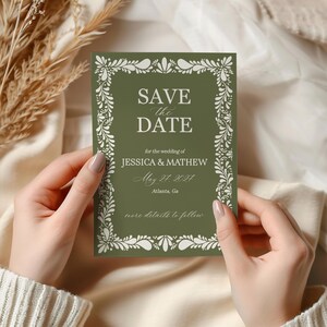 May include: A rectangular olive green "Save the Date" card with white floral border and text. The card reads "SAVE the DATE" and includes wedding details for Jessica & Mathew, May 24, 2024, Atlanta, GA. The card is held by two hands.