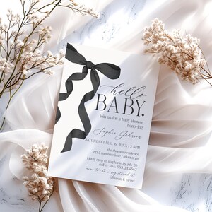 May include: A baby shower invitation with the words "hello BABY" in elegant script. A black ribbon adorns the top left corner, with the invitation details below. The invitation includes the date, time, location, and RSVP information.