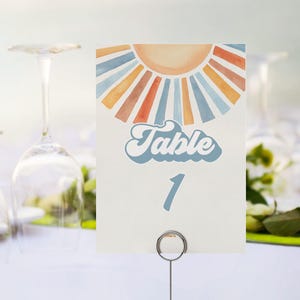 May include: A table number card with a retro sunburst design in orange, blue, and yellow. The word "Table" is written in a script font, with the number "1" below. The card is held by a silver metal holder.