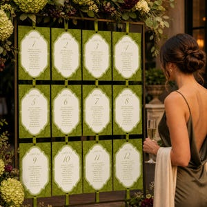 May include: A wedding seating chart display featuring twelve individual white cards with ornate borders, each labeled with a table number from one to twelve, mounted on a dark metal frame and decorated with green and purple floral arrangements.