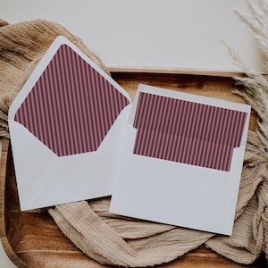 May include: Two white envelopes with burgundy and pink striped interior lining, one open to reveal the pattern. The envelopes and card are on a wooden tray with a tan fabric and dried plants.