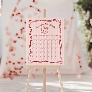 May include: A pink and white striped sign on a wooden easel. The sign reads "Guess the Date" and includes a calendar grid. The text "When will baby Madison arrive?" is also present. Decorative hearts are scattered around the sign.