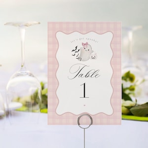 May include: A rectangular table number card featuring a light pink gingham background and a scalloped white center panel. The card displays the text "let's get spooky!" and "Table 1" with a small ghost illustration wearing a pink bow and bats.