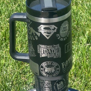 Peut inclure: Mug de voyage isotherme noir avec une poignée, avec le mot "Superdad" et diverses phrases liées aux pères. Le mug est posé sur une surface blanche. Idéal pour les boissons chaudes ou froides.