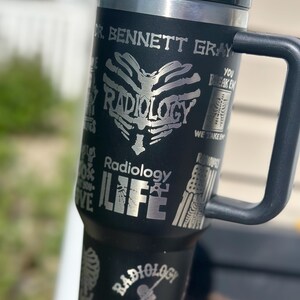 May include: Black travel mug with a silver design featuring the word "Radiology" and a rib cage heart. The mug has a black handle and lid. Text includes "Dr. Bennett Gray" and "Radiology Life".