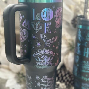 Könnte beinhalten: Schwarzer Reisebecher mit Griff, mit schillernden Akzenten und verschiedenen Grafiken. Der Becher ist mit dem Wort "LOVE", einer Eule und dem Text "Ollivander's Wands" verziert. Der Becher hat einen schwarzen Deckel und einen schwarzen Griff.