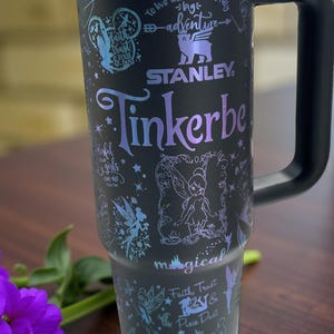 Puede incluir: Vaso Stanley negro con asa, con un diseño de Campanilla. El vaso tiene un asa negra y un borde plateado. El diseño incluye a Campanilla, estrellas y las palabras "Faith, Trust & Pixie Dust". También es visible el texto "To live will be an awfully big adventure".