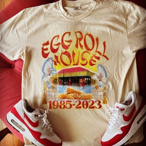 May include: Cream-colored t-shirt with a graphic that reads "EGG ROLL HOUSE 1985-2023" in red and yellow. The graphic includes an illustration of a building and two white doves. A pair of white and red sneakers are placed on either side of the shirt.