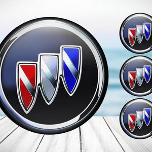 Może przedstawiać: Trzy srebrne, chromowane emblematy z czerwonymi, białymi i niebieskimi akcentami. Emblematy są ułożone w kształcie trójkąta i umieszczone na czarnym tle.