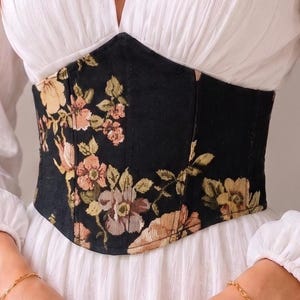 Puede incluir: Un vestido blanco de manga larga con un escote en V profundo y un panel de cintura estructurado estilo corsé negro. El corsé está decorado con un estampado floral en tonos rosa, amarillo y verde. El vestido tiene puños fruncidos y una falda fluida.