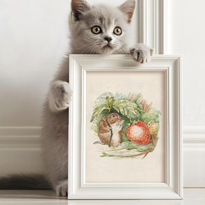 Pode incluir: Um gato cinza espreita por cima de uma moldura branca com uma ilustração em aquarela de um rato marrom comendo um morango. A ilustração está sobre um fundo creme.